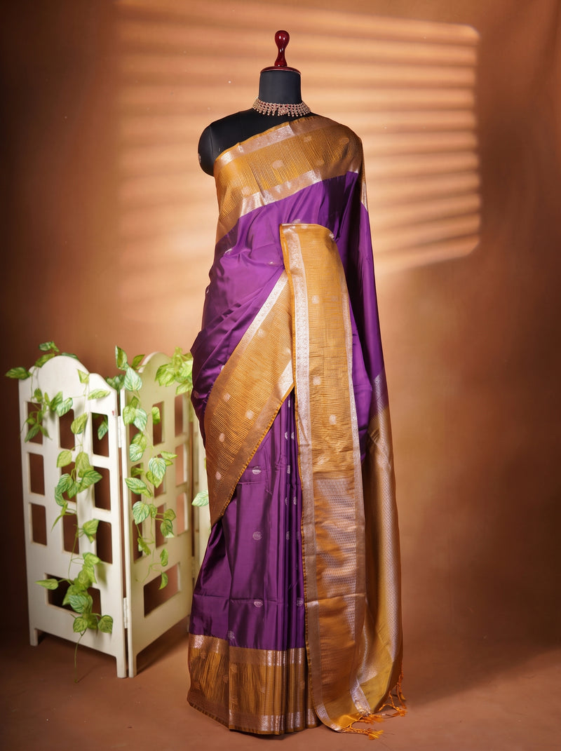 A purple silk saree with a metallic gold border and pallu draped on a black mannequin, set against a warm brown backdrop with a decorative white planter on the left.