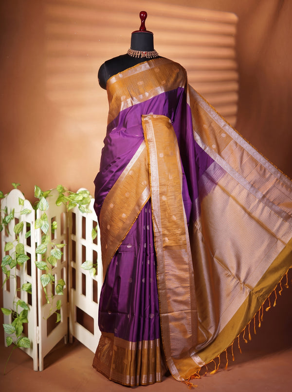 A purple silk saree with a wide gold zari border and pallu, draped on a black mannequin torso. The saree is styled with a matching purple and gold blouse, set against a warm brown background with a decorative white screen and green foliage.