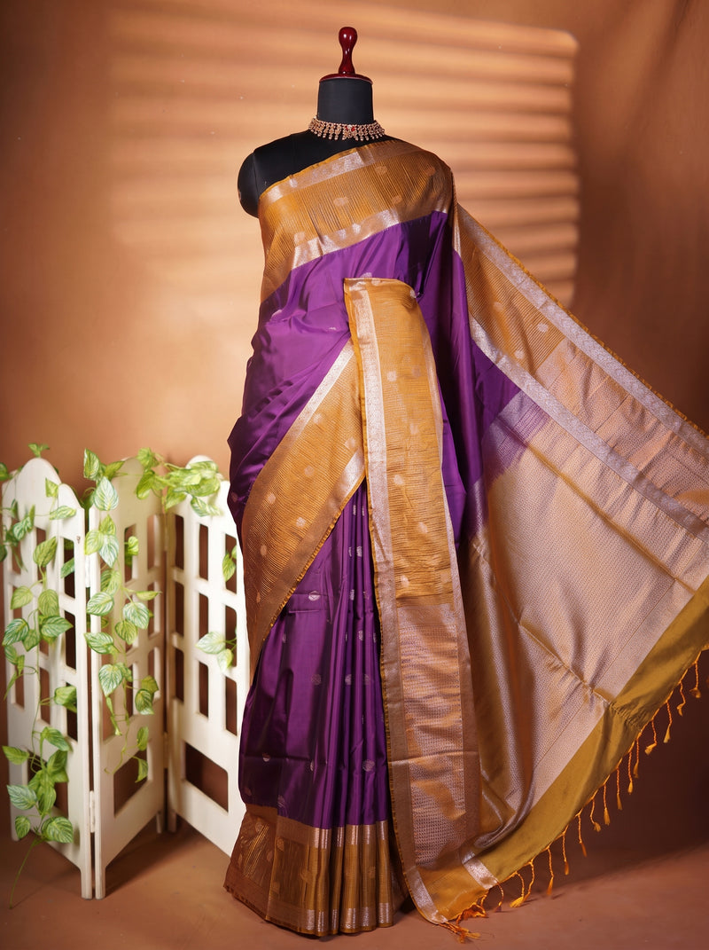 A purple silk saree with a wide gold zari border and pallu, draped on a black mannequin torso. The saree is styled with a matching purple and gold blouse, set against a warm brown background with a decorative white screen and green foliage.