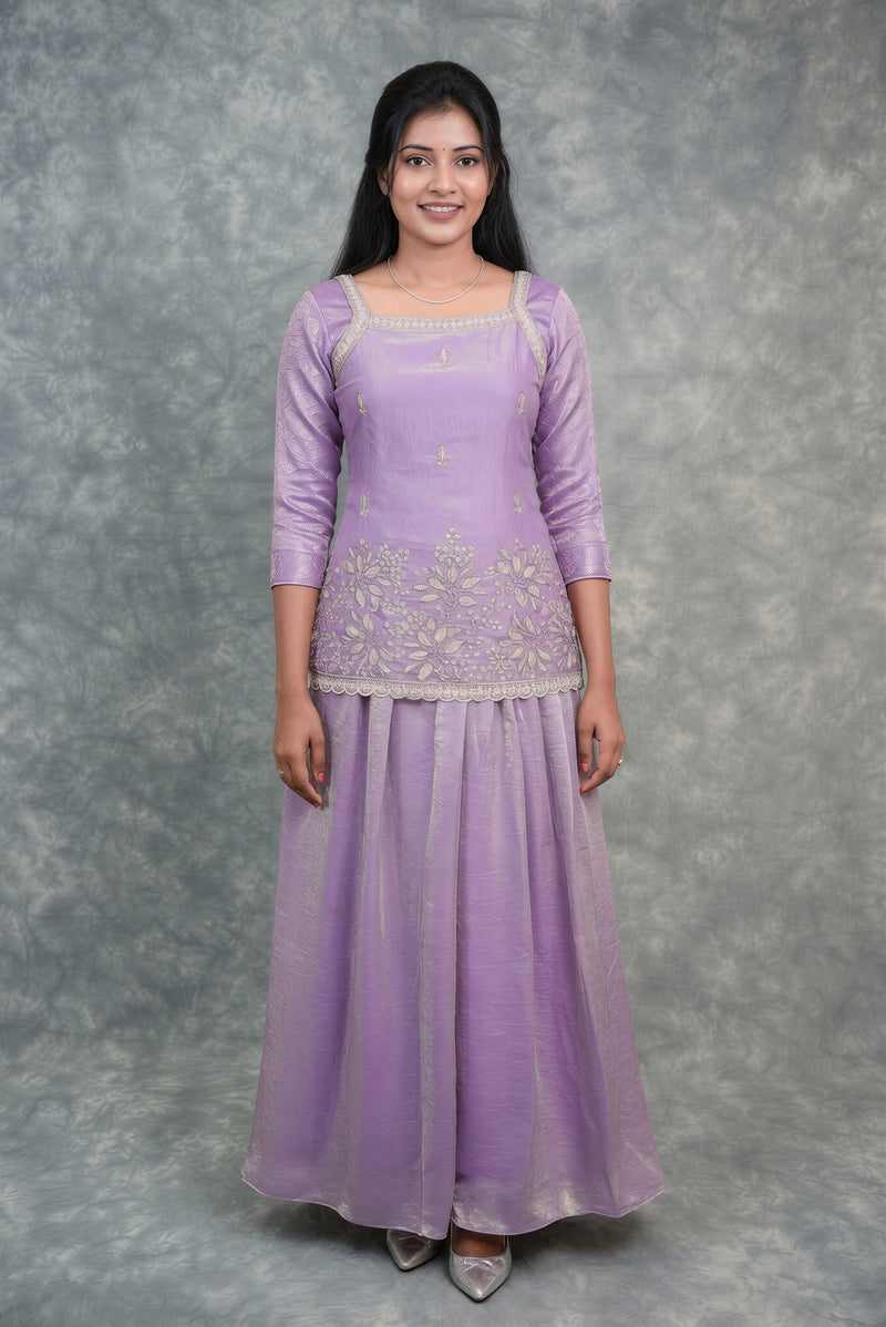 A woman modeling a lavender purple traditional Indian outfit consisting of a long-sleeved embroidered kurta and a matching flared skirt, standing against a mottled gray studio backdrop.