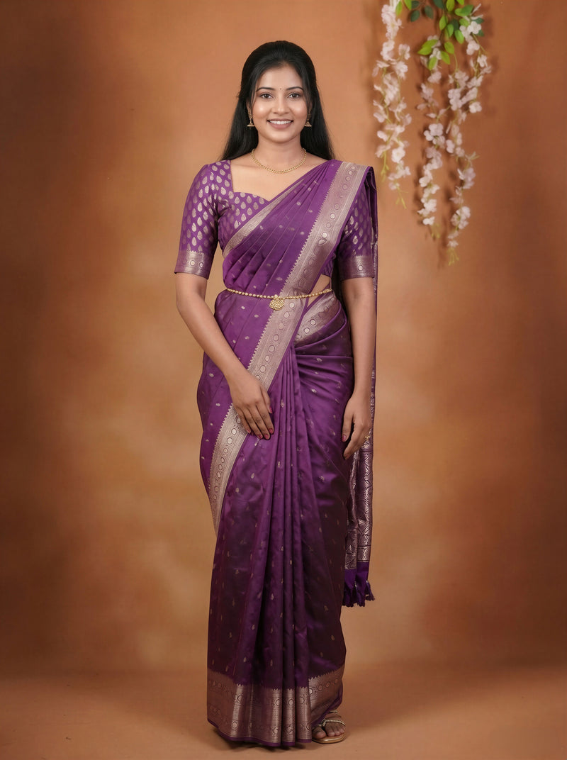 A woman modeling a purple silk saree with gold zari patterns and borders, standing against a warm brown studio backdrop with hanging floral decorations.