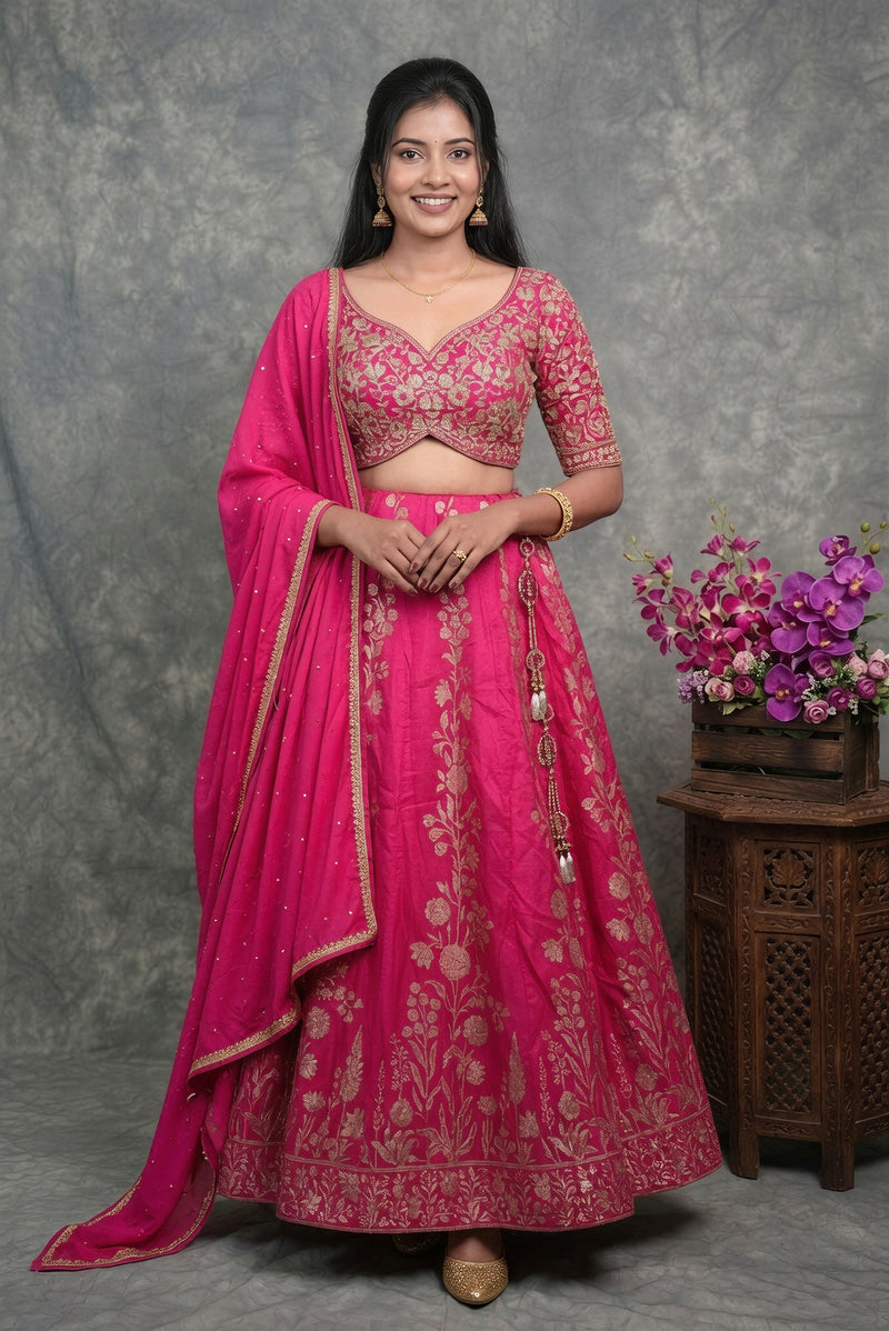 A woman modeling a bright pink traditional Indian lehenga choli set with intricate gold embroidery, standing against a textured gray backdrop next to a wooden table with purple flowers.