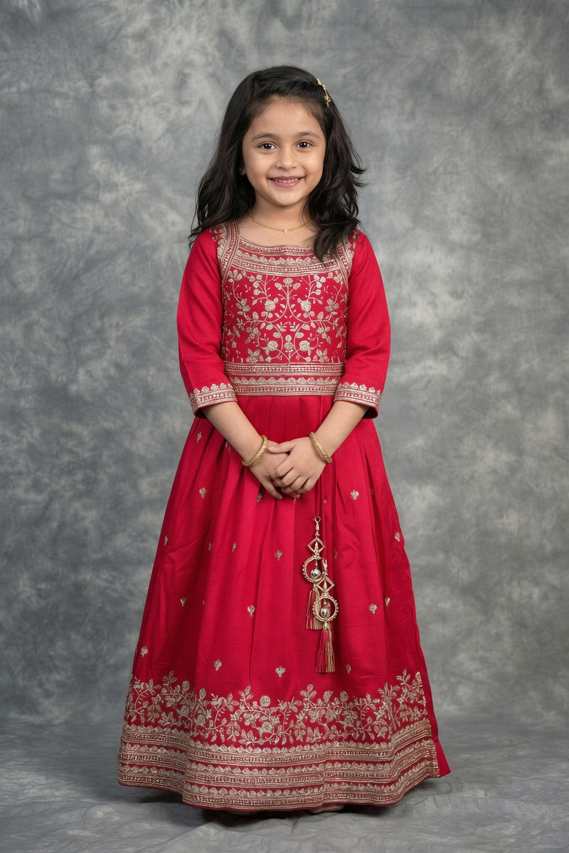 A young girl modeling a bright red traditional Anarkali gown with intricate gold and silver embroidery, standing against a textured gray studio backdrop.