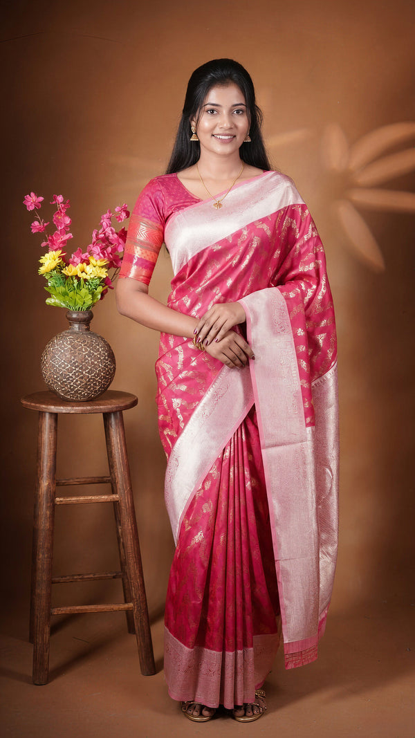 A woman wearing a pink and white silk saree stands in front of a brown studio backdrop, with a wooden stool and vase of flowers to her left.