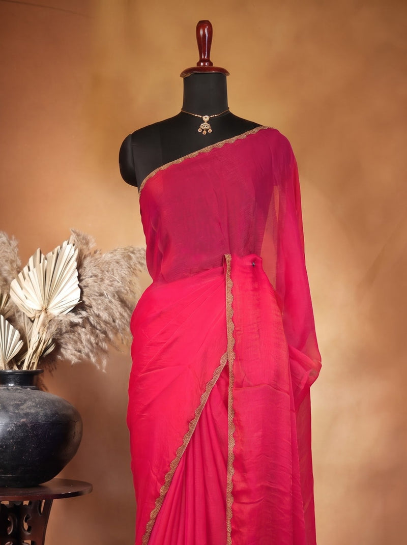 A bright pinkish-red saree displayed on a black mannequin dress form, styled with a matching blouse. The set is photographed against a warm brown studio backdrop, with a decorative dark vase and dried palm leaves on a carved wooden stand to the left.