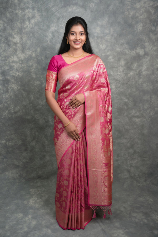 A woman modeling a pink and rose gold saree with gold zari patterns, standing against a textured gray studio backdrop.