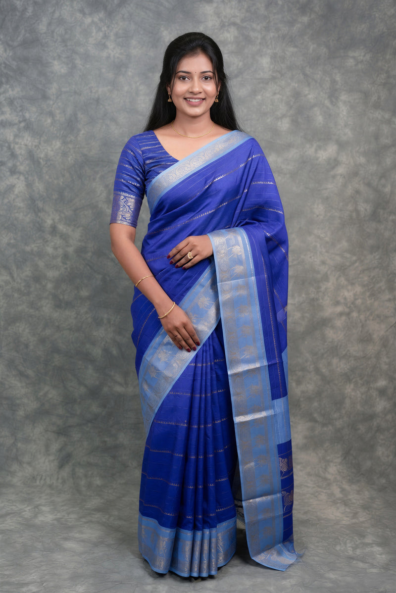 A woman modeling a royal blue and light blue saree with a matching blue blouse, standing against a textured gray studio backdrop.