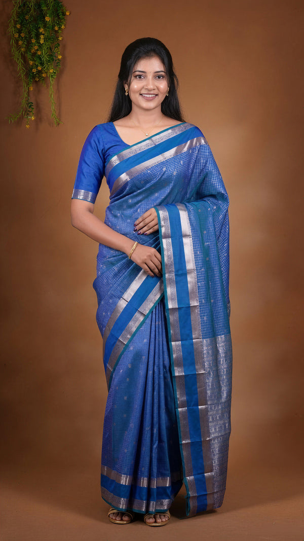 A woman modeling a royal blue silk saree with silver zari stripes and a matching blue blouse, set against a solid brown studio backdrop.