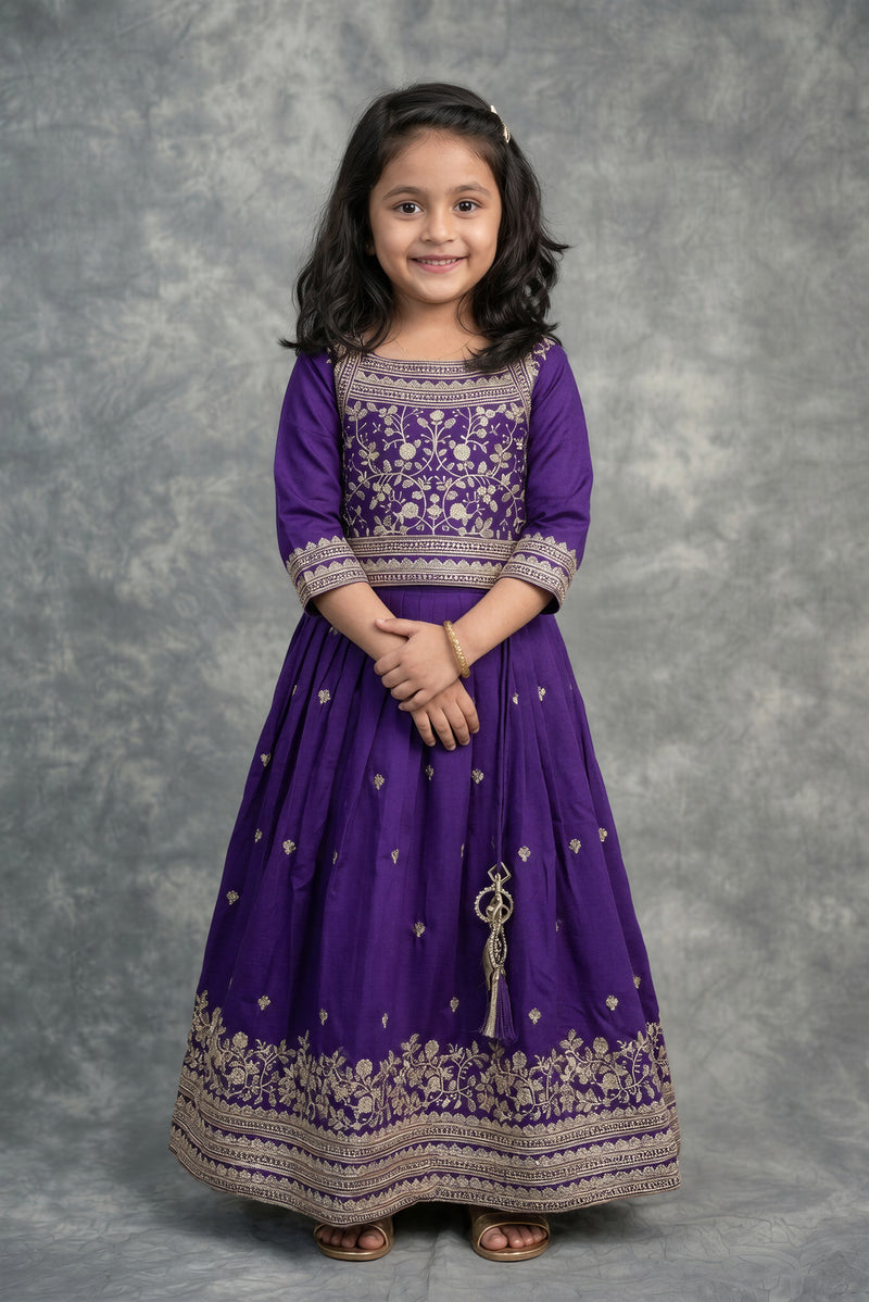 A young girl with dark hair wearing a purple traditional ethnic dress with intricate gold embroidery, standing against a textured gray studio backdrop.