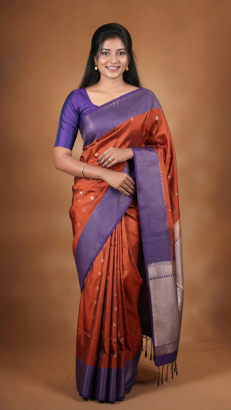 A woman modeling a traditional Indian saree with a purple blouse and a rust-orange saree body with a purple border and pallu, set against a solid brown studio background.