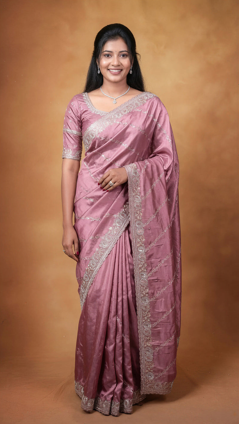 A woman modeling a dusty pink silk saree with silver embroidered borders and a matching embroidered blouse, set against a warm brown studio backdrop.