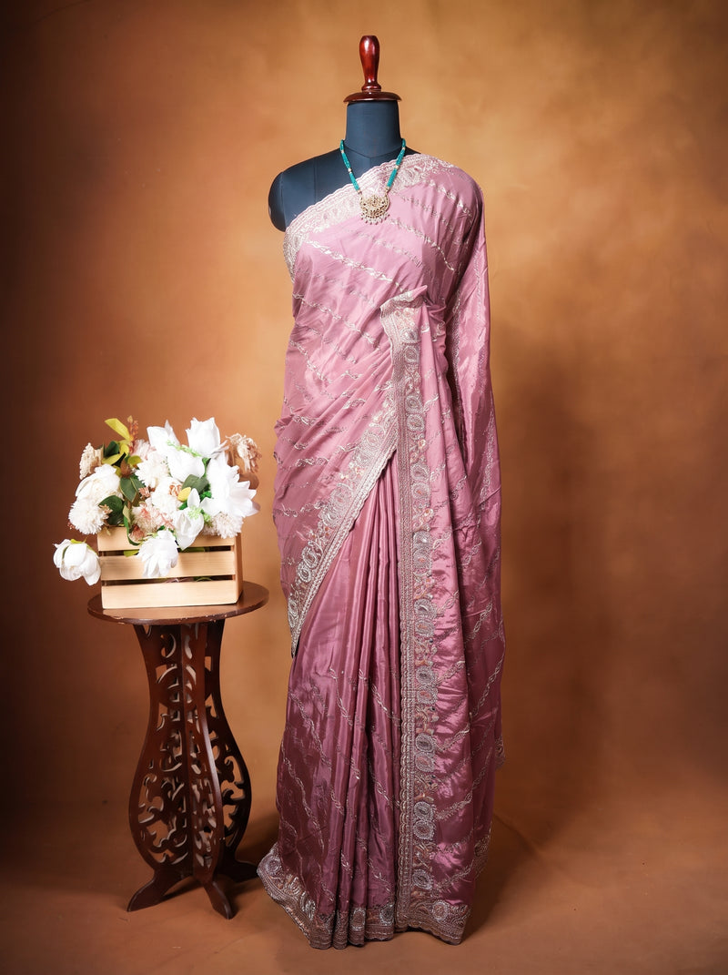 A mauve pink saree with silver embroidery displayed on a black mannequin torso, set against a warm brown studio backdrop. A small wooden side table with white flowers is positioned to the left of the mannequin.