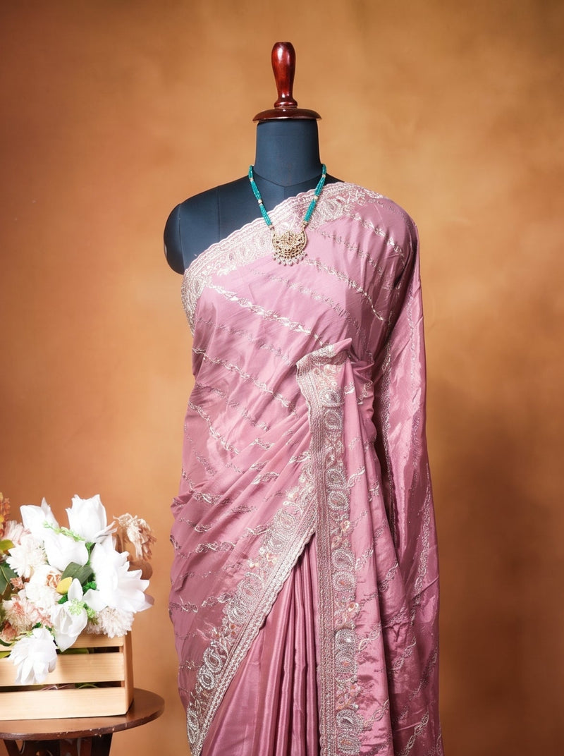 A mauve pink saree with silver embroidery displayed on a black mannequin torso, set against a warm brown studio backdrop. A small wooden side table with white flowers is positioned to the left of the mannequin.