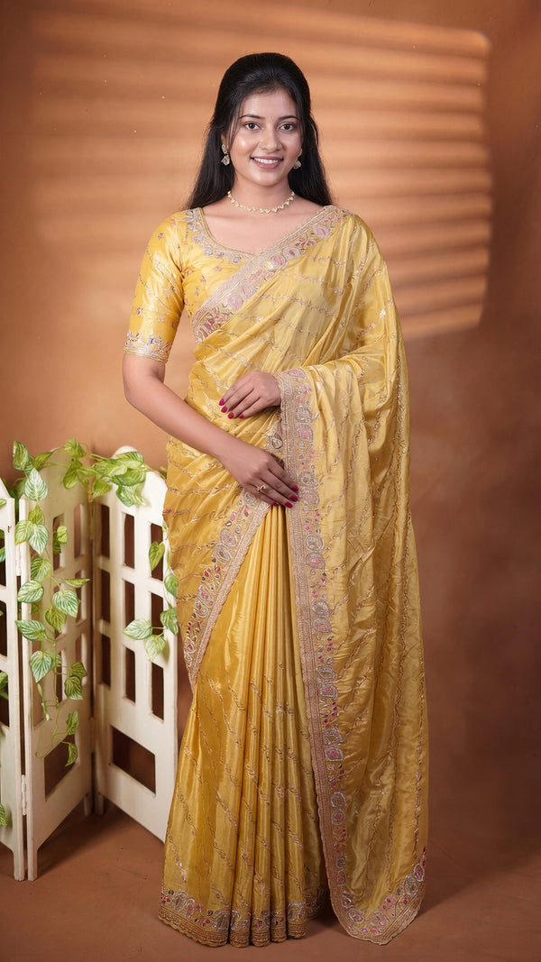 A woman modeling a yellow saree with intricate silver and gold border embroidery, standing against a warm brown backdrop with a decorative white screen and green leaves on the left.