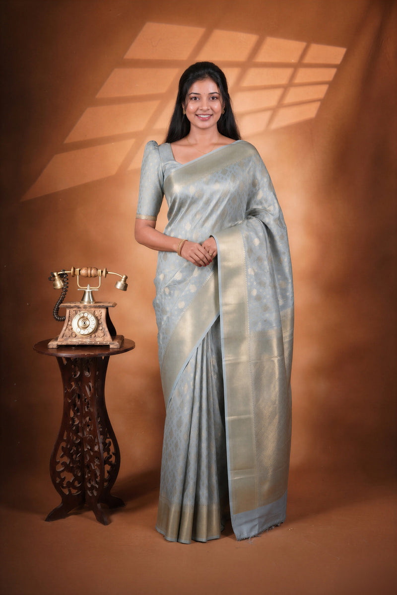 A woman wearing a light blue saree with gold zari embroidery stands next to a vintage wooden telephone on a pedestal against a warm brown studio backdrop.