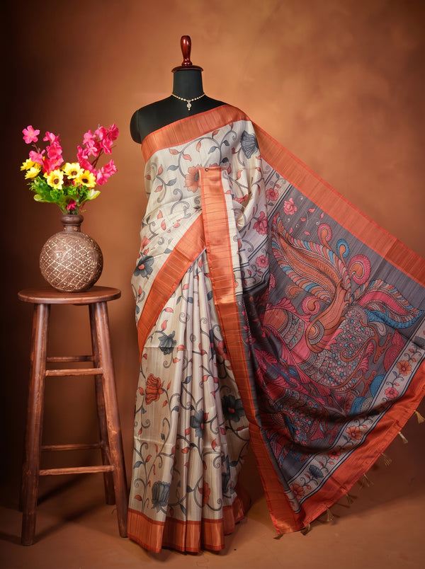 A printed cotton saree displayed on a black mannequin torso, styled with a decorative wooden stool holding a vase of colorful flowers against a warm brown studio backdrop. The saree has a light beige base with blue, red, and orange floral motifs and a wide orange border.