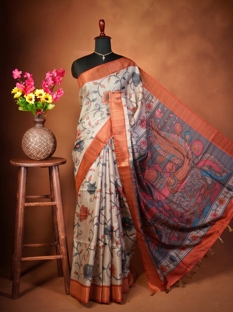 A printed cotton saree displayed on a black mannequin torso, styled with a decorative wooden stool holding a vase of colorful flowers against a warm brown studio backdrop. The saree has a light beige base with blue, red, and orange floral motifs and a wide orange border.