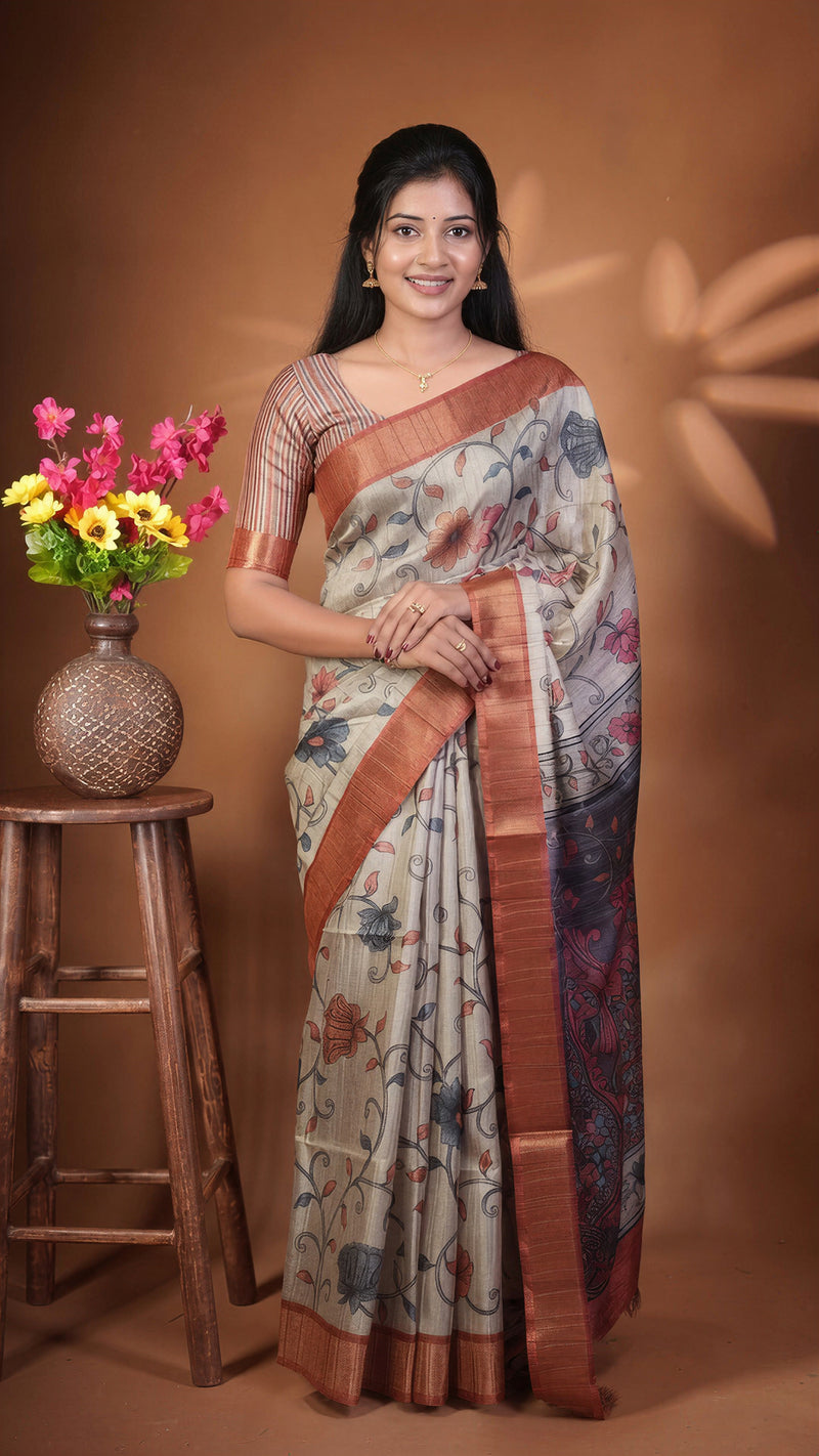 A woman models a traditional Indian saree with a beige base, brown border, and floral patterns, standing next to a wooden stool with a vase of flowers against a warm brown backdrop.
