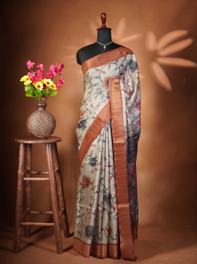 A patterned saree displayed on a black mannequin torso, styled next to a wooden stool holding a vase of pink and yellow flowers against a warm brown backdrop.