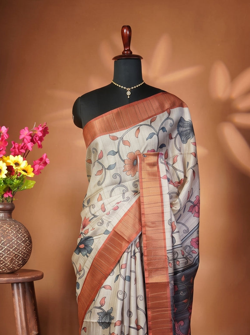 A patterned saree displayed on a black mannequin torso, styled next to a wooden stool holding a vase of pink and yellow flowers against a warm brown backdrop.