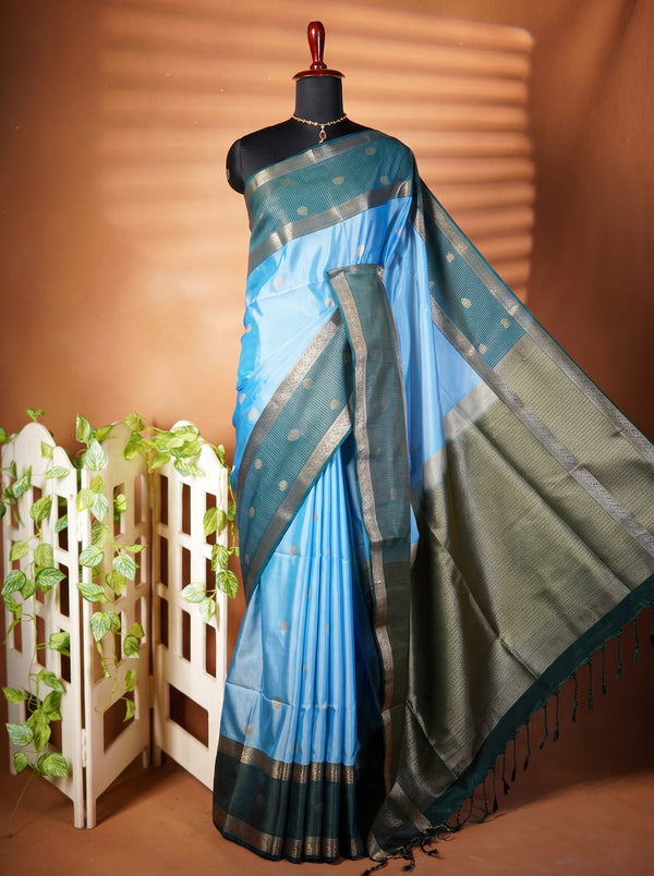 A blue and green saree displayed on a black mannequin, featuring a light blue body, a dark green border, and metallic gold zari patterns, set against a warm brown background with a decorative white screen.