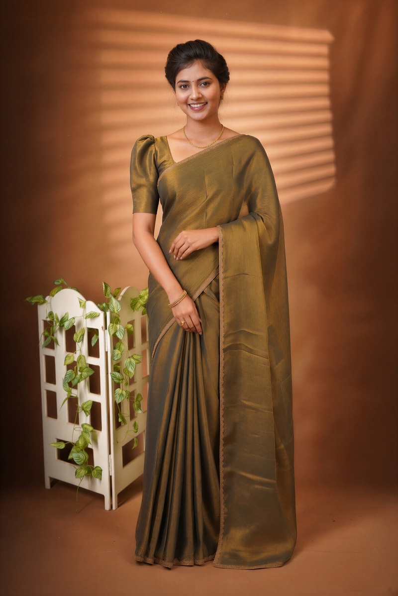 A woman wearing a draped olive green saree stands against a warm brown studio backdrop with soft window-blind light shadows. A small white decorative stand with green vines is beside her.