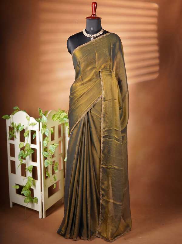A draped olive green saree displayed on a black mannequin with a red top, styled with a pearl necklace. The mannequin is set against a warm brown backdrop with a decorative white screen and green leaves on the left.