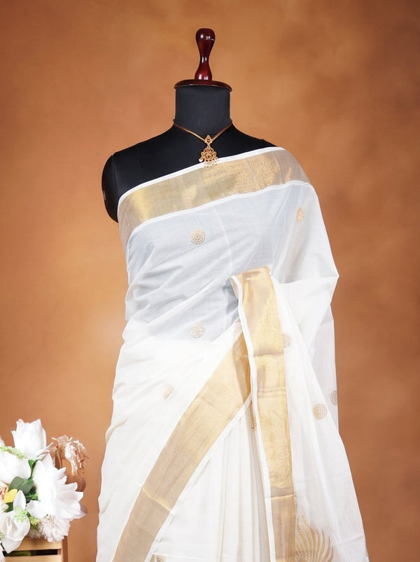 A white silk saree with gold borders and motifs displayed on a black mannequin, next to a small wooden table holding a bouquet of white flowers, against a warm brown studio backdrop.