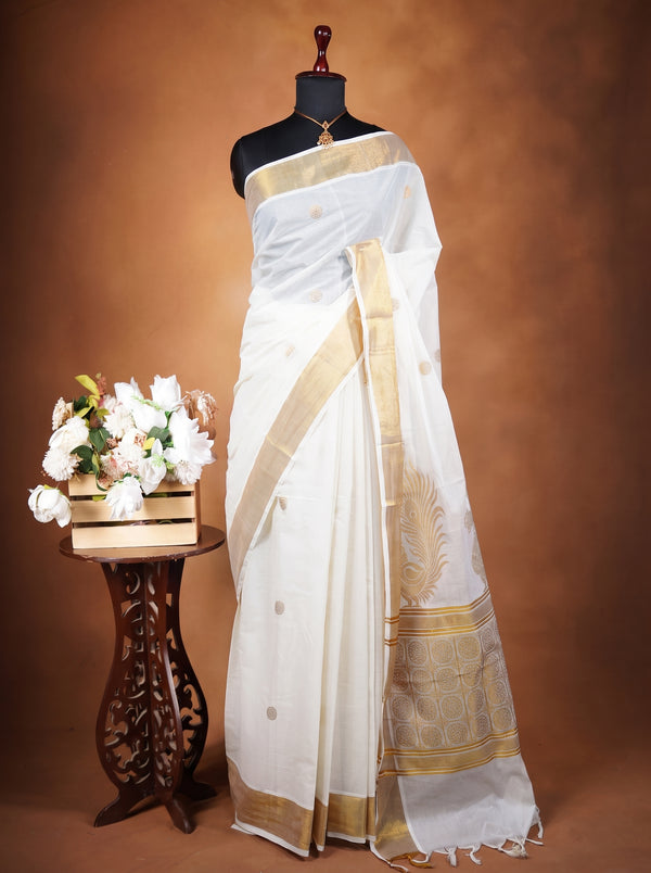 A white silk saree with gold borders and motifs displayed on a black mannequin, next to a small wooden table holding a bouquet of white flowers, against a warm brown studio backdrop.