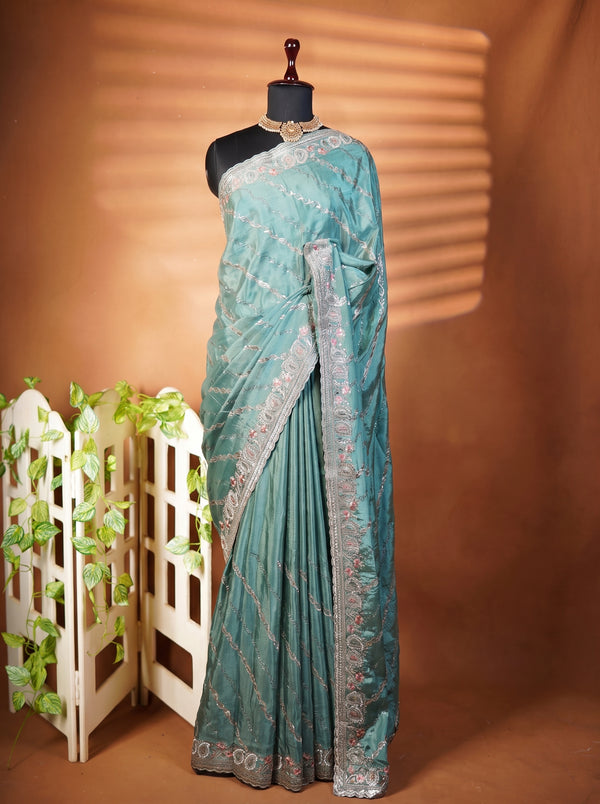 A teal blue-green saree with silver floral embroidery and a matching border, displayed on a black mannequin torso. The mannequin is set against a warm brown backdrop with soft horizontal shadows, and a small white decorative screen with green vines is visible on the left.