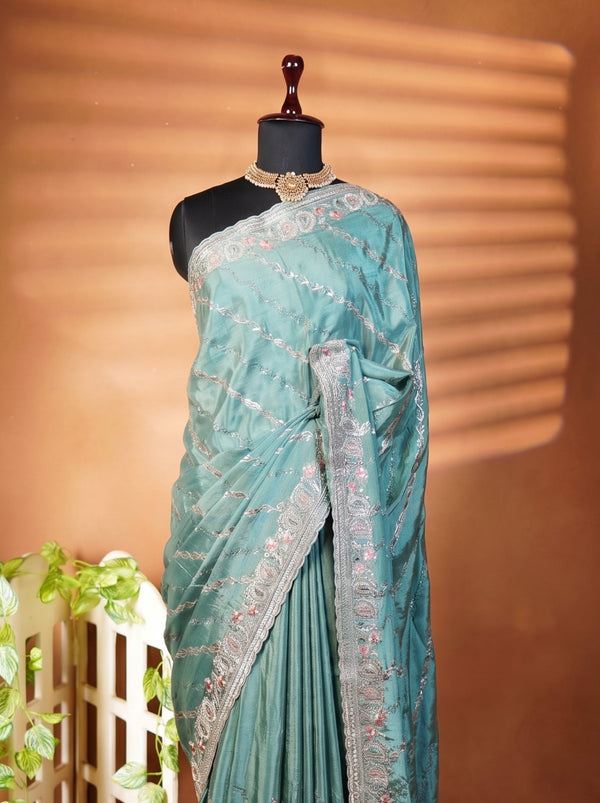 A teal blue-green saree with silver floral embroidery and a matching border, displayed on a black mannequin torso. The mannequin is set against a warm brown backdrop with soft horizontal shadows, and a small white decorative screen with green vines is visible on the left.