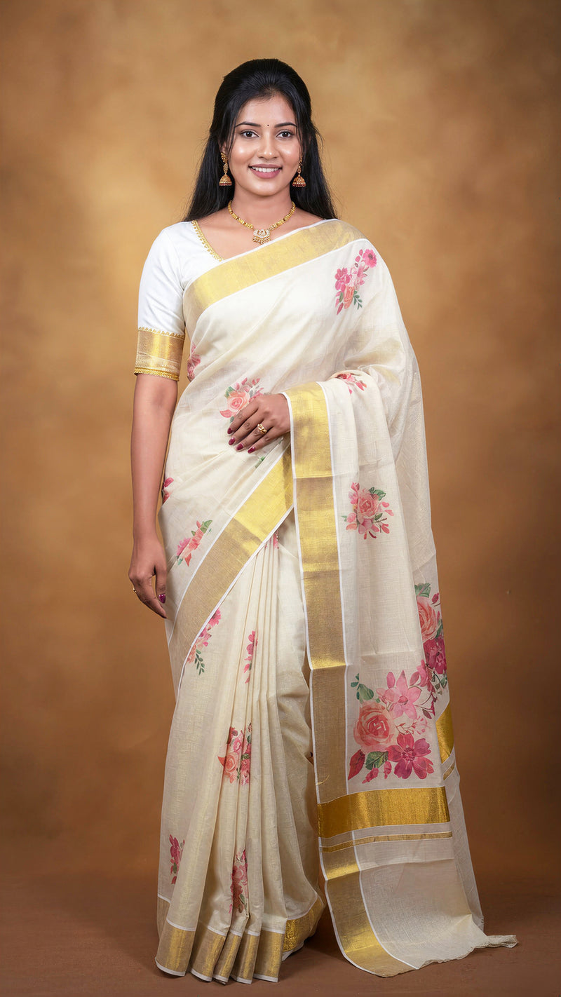 A woman modeling a cream-colored silk saree with gold borders and pink floral prints, paired with a white short-sleeve blouse, set against a warm brown studio backdrop.