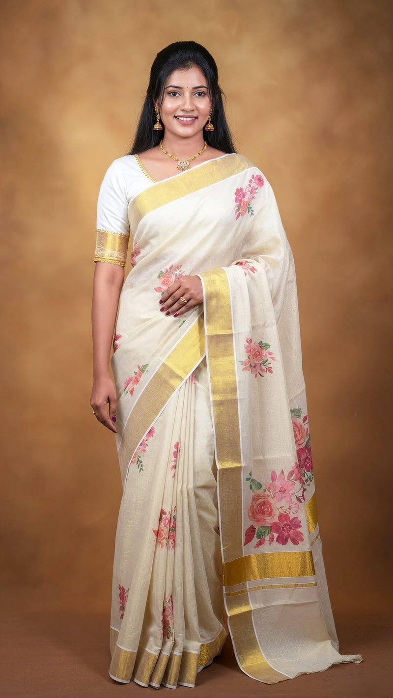 A woman modeling a cream-colored silk saree with gold borders and pink floral prints, paired with a white short-sleeve blouse, set against a warm brown studio backdrop.