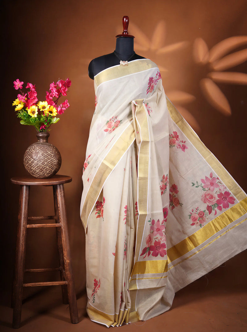 A cream-colored cotton saree with pink floral prints and gold borders, draped on a black mannequin torso. The mannequin is set against a warm brown background with decorative leaf shadows, and a small wooden stool with a vase of colorful flowers is on the left.