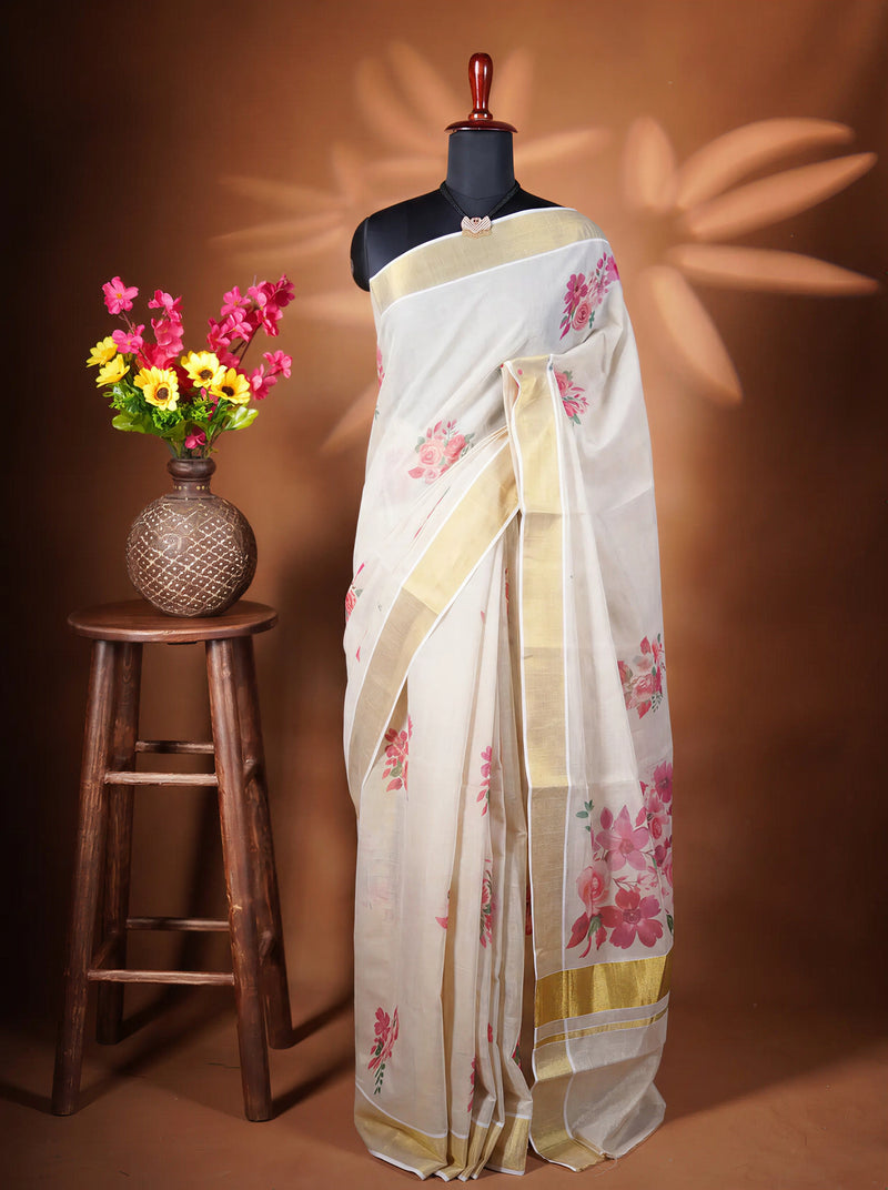 A white saree with pink floral prints and gold borders displayed on a black mannequin torso, next to a wooden stool holding a vase of colorful flowers against a warm brown backdrop.