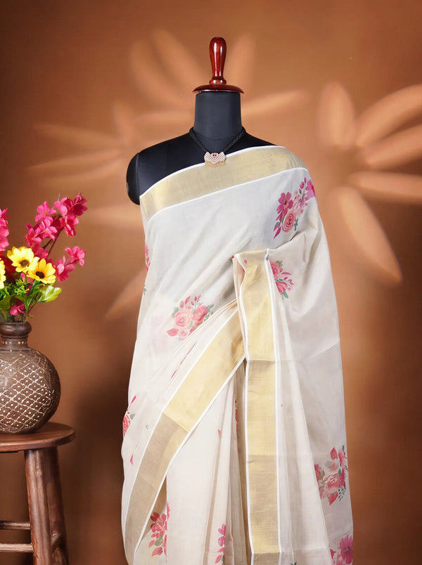 A white saree with pink floral prints and gold borders displayed on a black mannequin torso, next to a wooden stool holding a vase of colorful flowers against a warm brown backdrop.