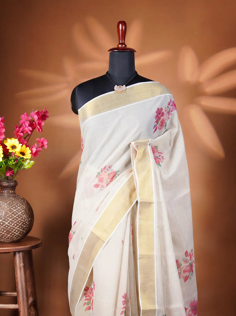 A white saree with pink floral prints and gold borders displayed on a black mannequin torso, next to a wooden stool holding a vase of colorful flowers against a warm brown backdrop.