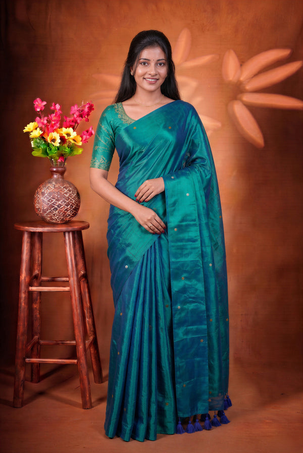 A woman wearing a teal green silk saree with gold zari embroidery and a matching embroidered blouse, standing in front of a warm brown backdrop with a wooden stool and flowers on the left.
