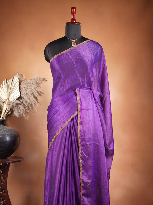 A purple silk saree with gold border detailing displayed on a black mannequin torso, set against a warm brown studio background with a decorative vase on the side.