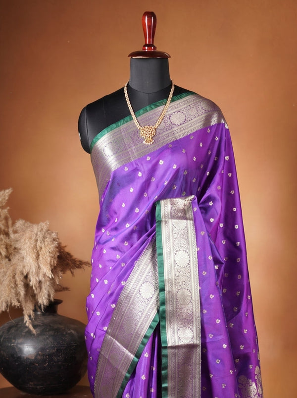 A purple and green silk saree with Gold zari embroidery, displayed on a black mannequin torso. The saree is styled with a matching green blouse, and the setup is photographed against a warm brown studio backdrop with a decorative wooden stand and vase on the left.