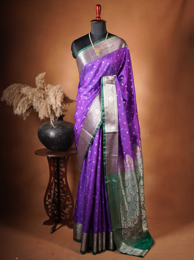 A purple and green silk saree with Gold zari embroidery, displayed on a black mannequin torso. The saree is styled with a matching green blouse, and the setup is photographed against a warm brown studio backdrop with a decorative wooden stand and vase on the left.