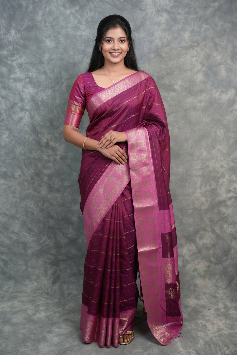 A woman modeling a traditional Indian silk saree in magenta purple with gold zari stripes and a matching blouse, standing against a textured gray backdrop.