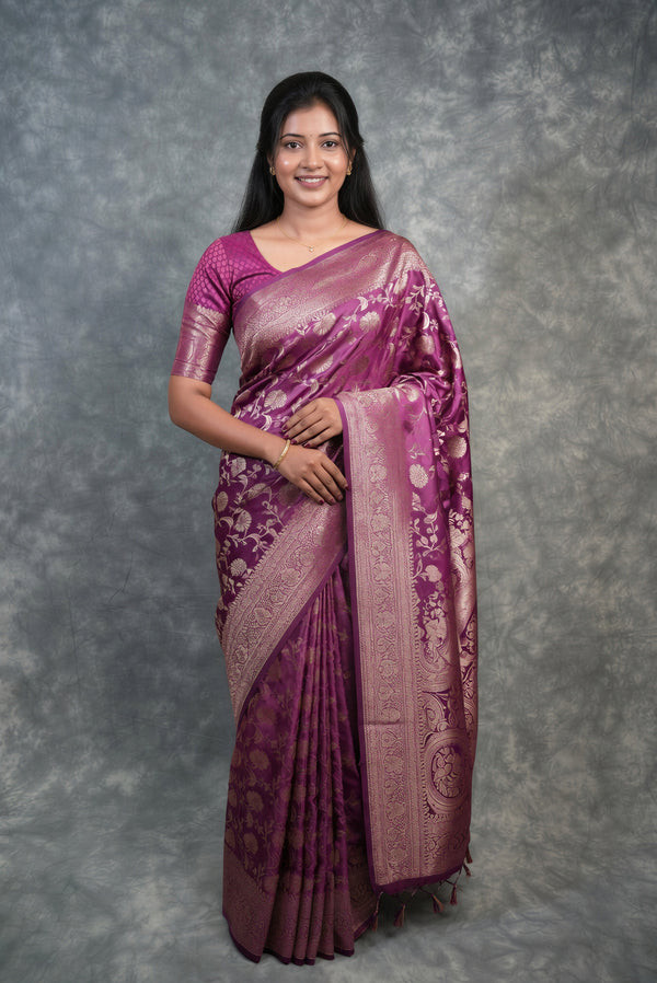 A woman modeling a purple and pink Banarasi silk saree with intricate gold zari floral patterns, standing against a textured gray studio backdrop.
