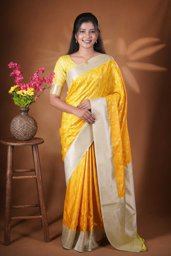 A woman modeling a yellow and beige silk saree with a matching printed blouse, standing next to a wooden stool with a flower vase against a brown studio backdrop.