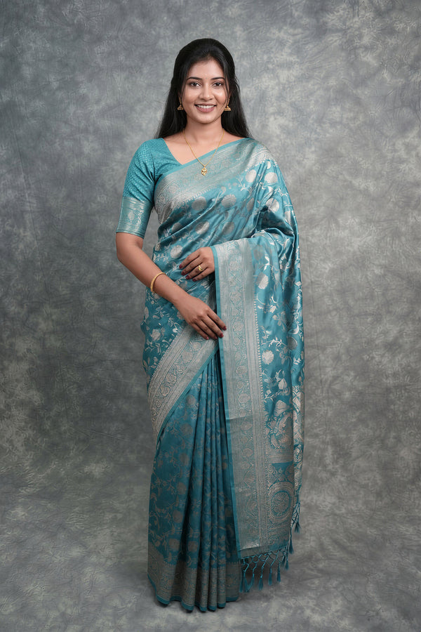 A woman modeling a teal blue and silver Patola-style saree with a matching blouse, standing against a textured gray studio backdrop.