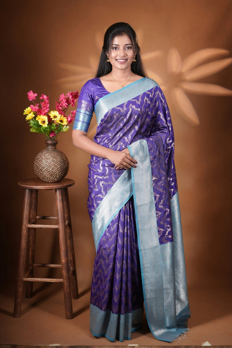 A woman wearing a purple and light blue patterned silk saree stands in front of a brown backdrop, next to a wooden stool holding a vase of colorful flowers.