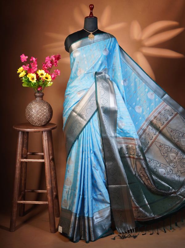 A light blue silk saree with a metallic silver border and traditional motifs, draped on a black mannequin torso. The mannequin is set against a warm brown background with leaf shadows, and a small wooden stool with a vase of colorful flowers is on the left.