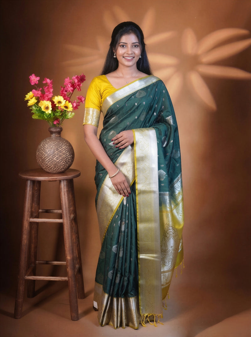 A woman wearing a dark green and gold saree with a yellow blouse, standing next to a wooden stool with a flower vase against a warm brown backdrop.