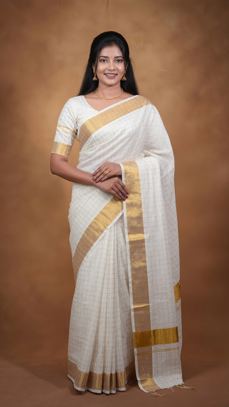 A woman modeling a white saree with a gold border and a subtle grid pattern, set against a solid brown studio backdrop.
