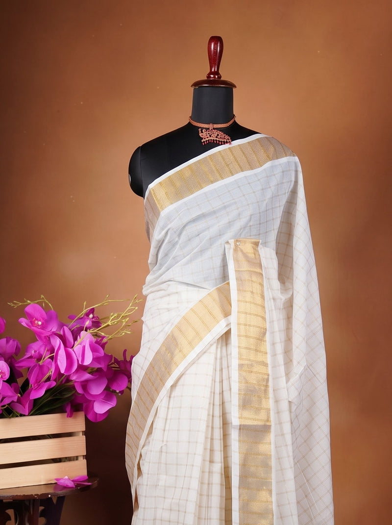 A white and gold saree draped on a black mannequin, styled next to a wooden crate filled with bright pink flowers on a decorative dark stand, set against a warm brown studio backdrop.